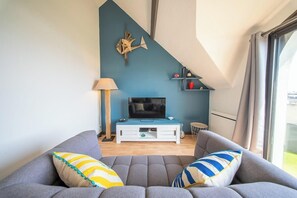 A modern living room with a grey sofa, a flat-screen TV on a white stand, a blue accent wall, and a window with curtains. - T2 avec aperçu mer, plage et commerces à pied (Larmor-Plage)