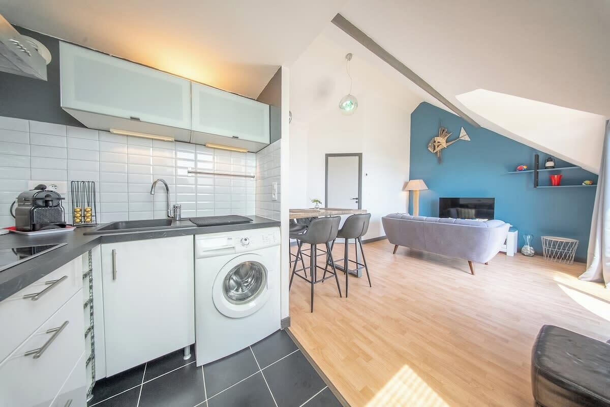 Une cuisine compacte équipée d’un lave-linge, un coin repas avec une table et des chaises, et un espace de vie comprenant un canapé et un téléviseur.