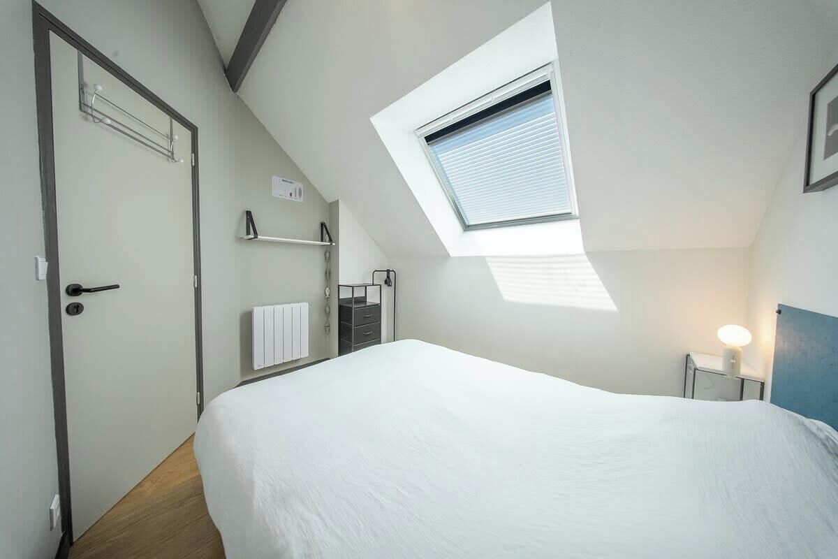 Une chambre à coucher avec un puits de lumière, un lit, une table de chevet avec une lampe, un radiateur et une porte.