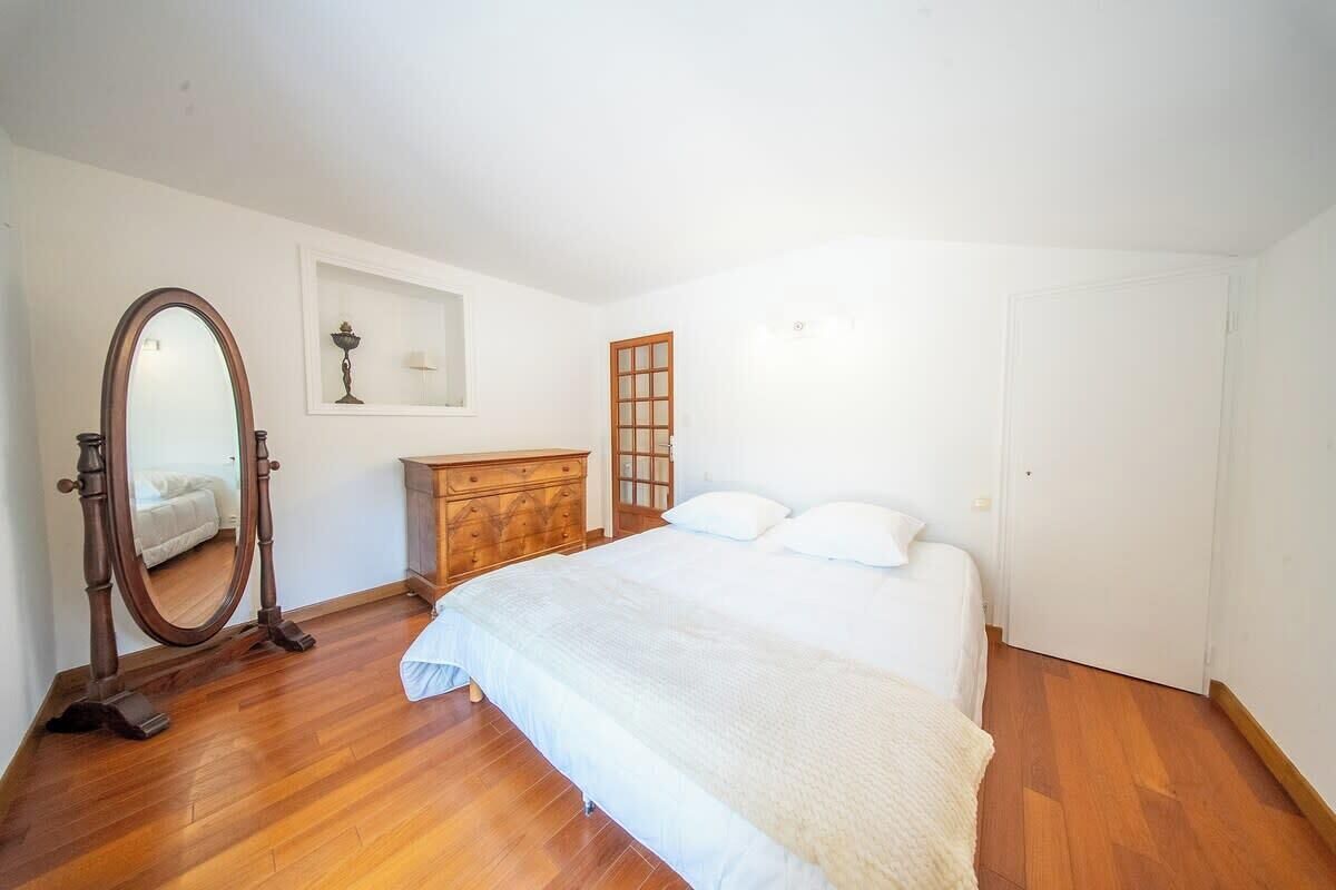 Een slaapkamer met een houten vloer, een bed met witte beddengoed, een dressoir, een spiegel en een deur.