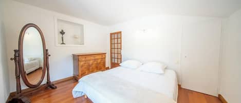 Un dormitorio con piso de madera, una cama con ropa de cama blanca, un tocador, un espejo y una puerta.