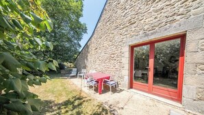 Un bâtiment en pierre doté d’une porte-fenêtre coulissante à cadre rouge, d’un patio avec une table et des chaises, et d’une zone herbeuse avec des arbres.
