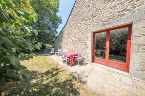 Ein Steinhaus mit roten Rahmelementen und einer Glasschiebetür, einer Terrasse mit Tisch und Stühlen und einem Rasen mit Bäumen.