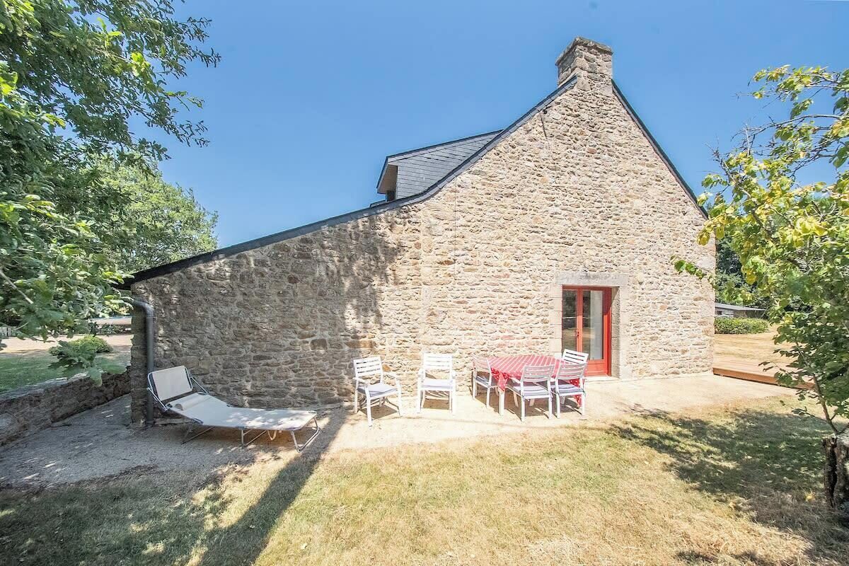 Un bâtiment en pierre avec une porte rouge, un petit patio avec une table et des chaises, et un fauteuil de relaxation.