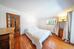 Ein Schlafzimmer mit einem hölzernen Schminktisch, einem Bett mit weißer Bettwäsche, einem Fenster mit Vorhängen und einem Heizkörper.