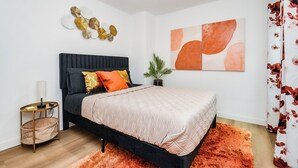Quarto moderno com cama preta, colcha acolchoada bege, mesa de cabeceira pequena com abajur, um objeto decorativo na parede e um vaso de planta.
