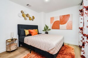 Ein modernes Schlafzimmer mit einem schwarzen Bett, einer beige gesteppten Tagesdecke, einem kleinen Nachttisch mit Lampe, einem Wanddekor und einem Blumentopf.