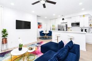 A modern living room with a sofa, coffee table, and a TV. There is a dining area with chairs and a table. The kitchen has white cabinets, a microwave, and a refrigerator. - Newly Built 3BR/3BA: Easy Access to Downtown, IAH, NRG, Med Center & Heights (Houston)