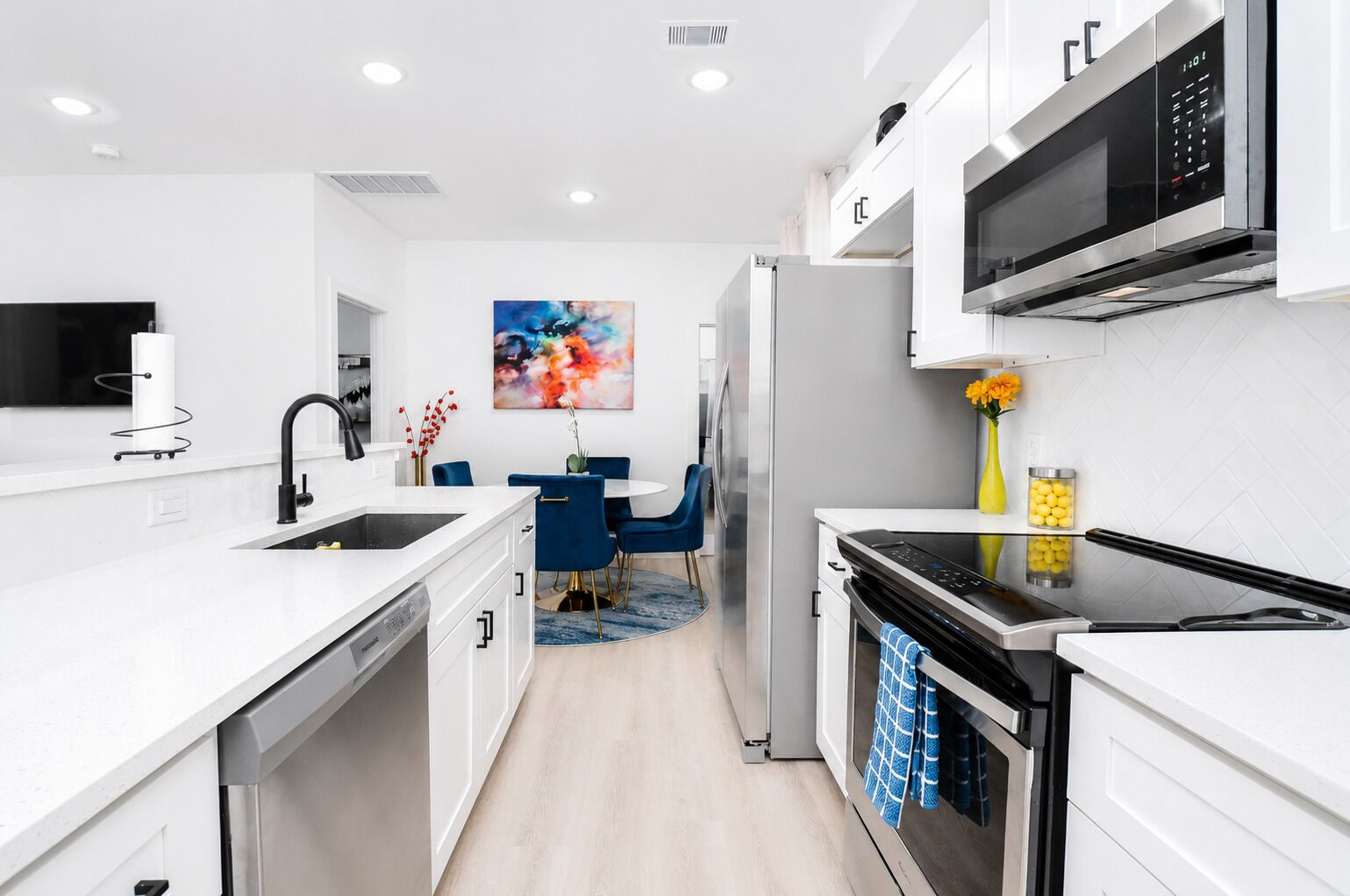 A modern kitchen with white cabinetry, a stainless steel refrigerator, a microwave, and a dining area with blue chairs.