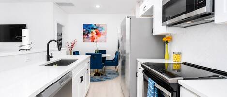 A modern kitchen with white cabinetry, a stainless steel refrigerator, a microwave, and a dining area with blue chairs.