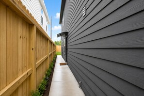 A narrow pathway flanked by wooden and grey siding walls, leading to a house with a visible door and window. - Newly Built 3BR/3BA: Easy Access to Downtown, IAH, NRG, Med Center & Heights (Houston)