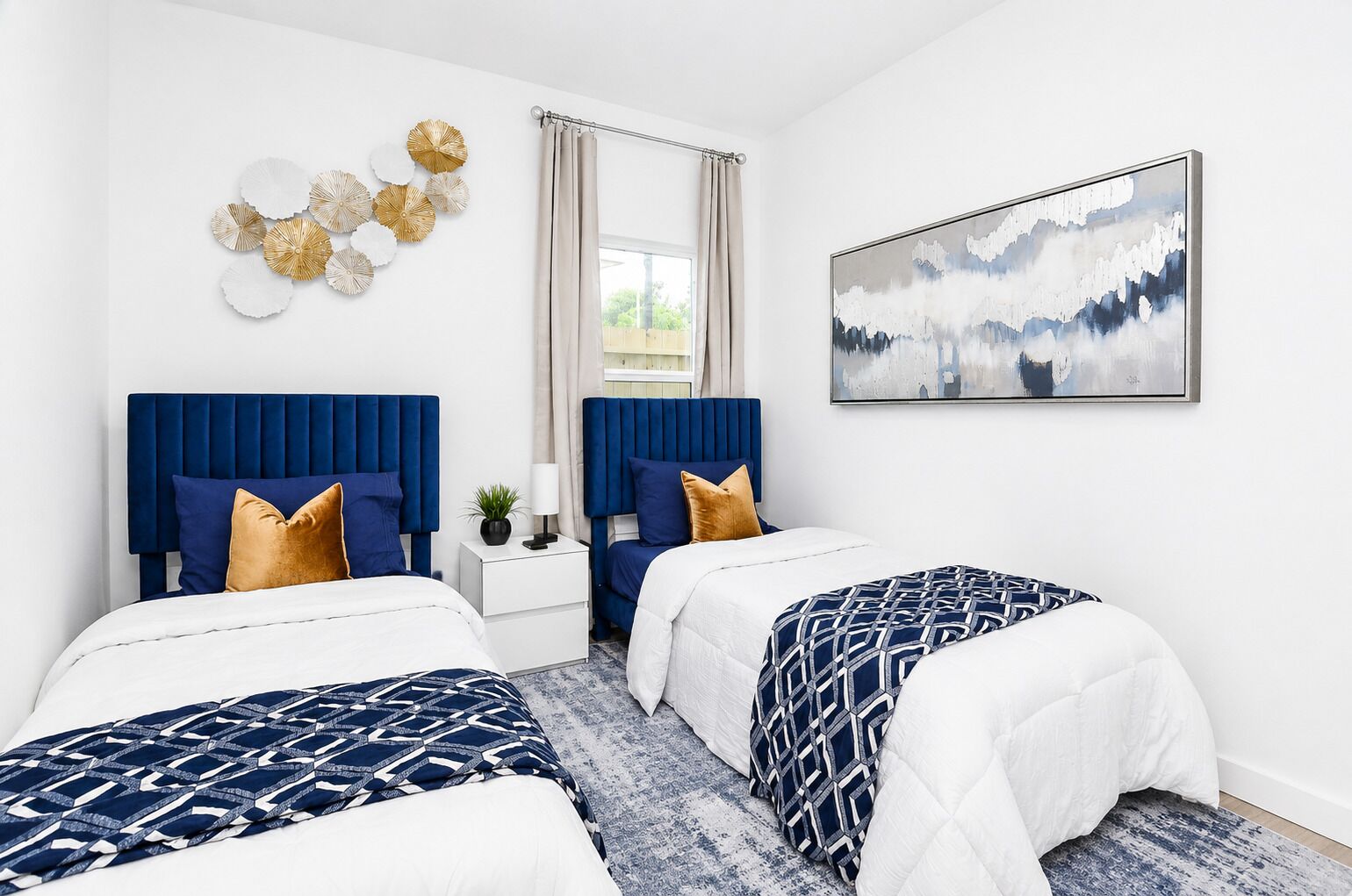 A hotel room with two beds, a nightstand, a window with curtains, a wall-mounted abstract painting, and a decorative wall piece.