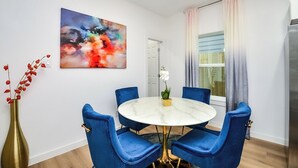 Uma sala de jantar com uma mesa redonda, cadeiras azuis, um vaso com flores vermelhas e um quadro abstrato colorido na parede.