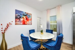 Ein Essbereich mit einem runden Tisch, blauen Stühlen, einer Vase mit roten Blumen und einem farbenfrohen, abstrakten Gemälde an der Wand.