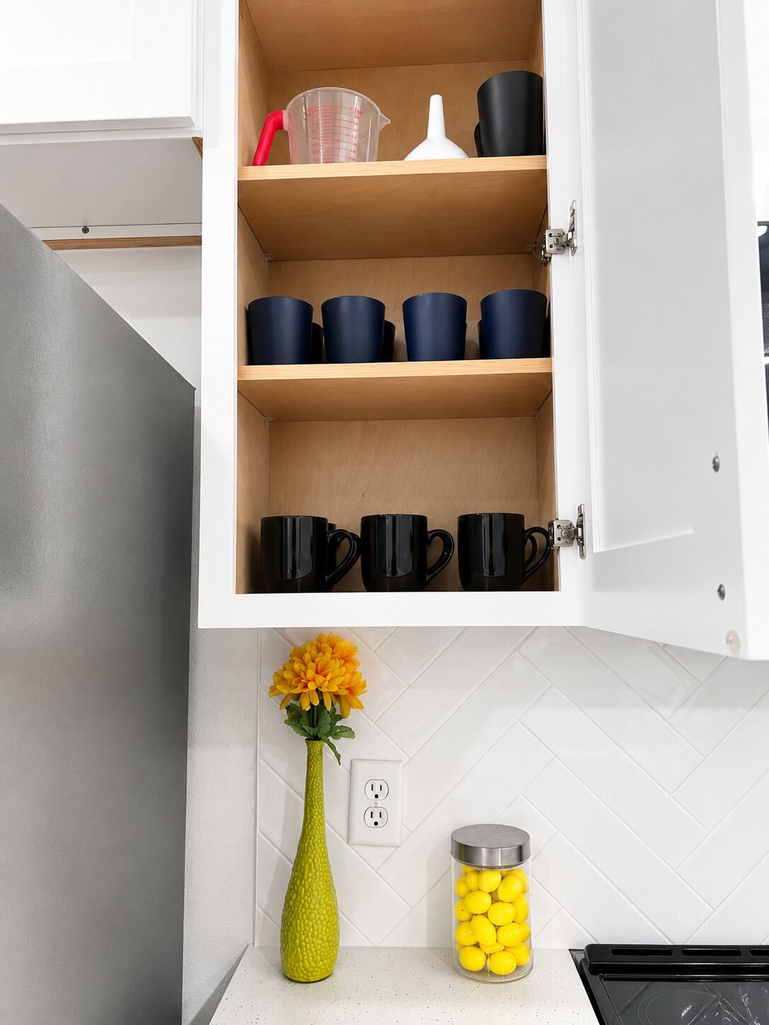 A kitchen with a cabinet containing black mugs, a measuring cup, and a white container.