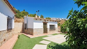 A white building with a stone base, a tiled roof, and a small garden area with a paved walkway.