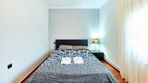 A single bed with a patterned bedspread, a bedside table with a lamp, and a window with curtains.