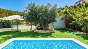 A swimming pool with a stone border, surrounded by artificial grass, trees, and a fence.