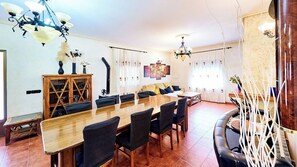 Comedor con una larga mesa de madera, sillas y un sofá. Hay dos lámparas de araña, un aparador con puertas de cristal y una pared con obras de arte enmarcadas.