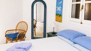 A bedroom with a bed, a chair, a nightstand, a lamp, a mirror, and a window with shutters.