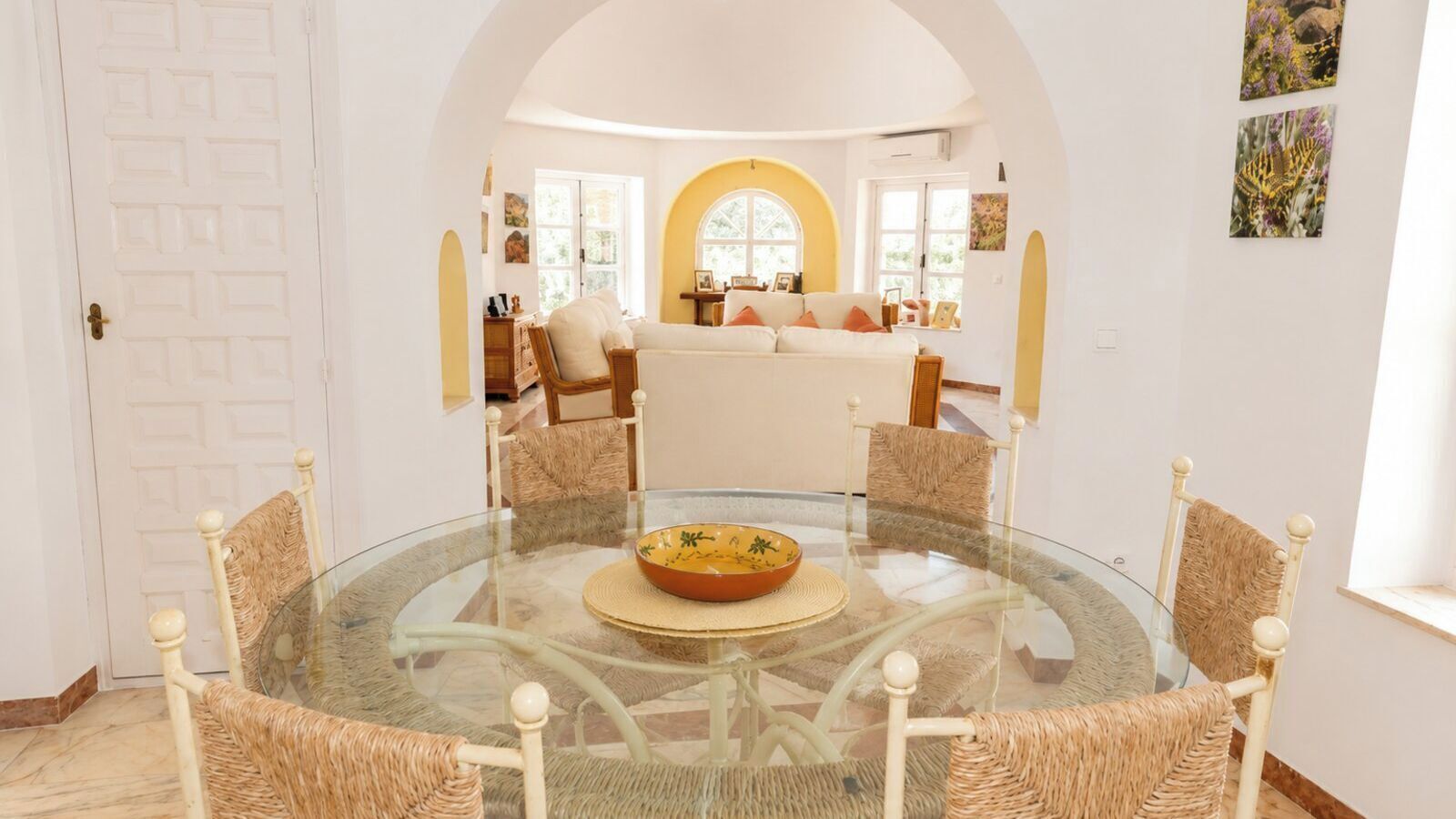 A dining area with a glass table surrounded by wicker chairs, a central bowl, and a view of a living room with a sofa and a window.