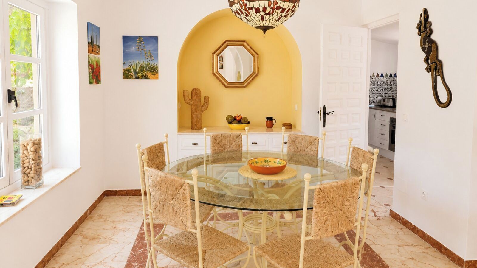 Un coin repas avec une table en verre entourée de chaises en osier, une décoration à base de cactus et un miroir au-dessus de la table.