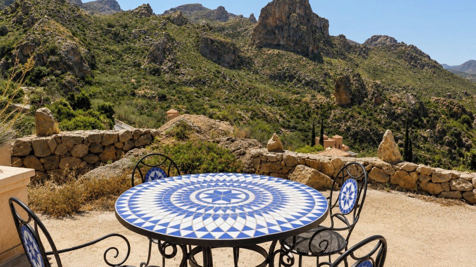An outdoor dining area with a patterned table and chairs, surrounded by a stone wall and a view of a mountainous landscape.