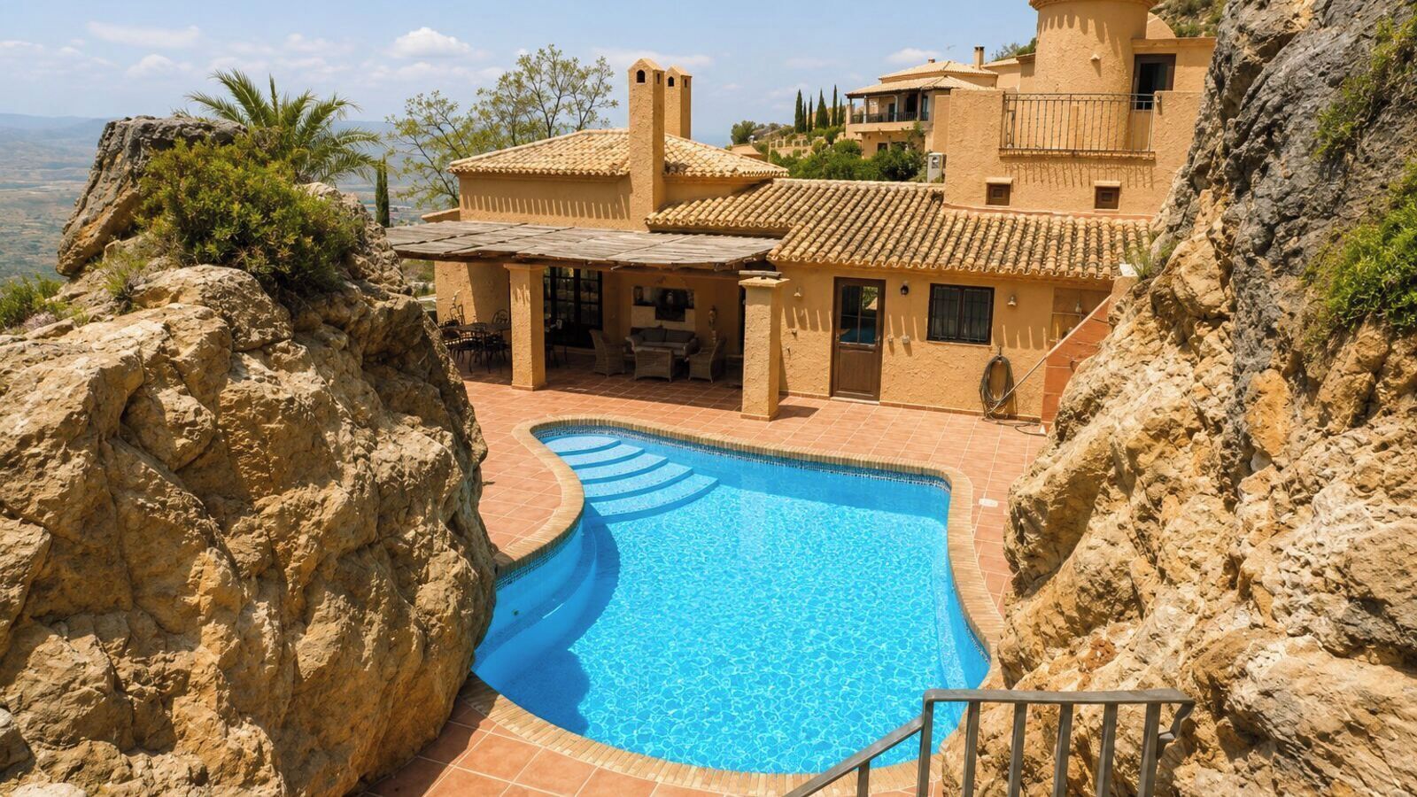 A pool with a curved edge, a patio area with outdoor furniture, and a building with a tiled roof.