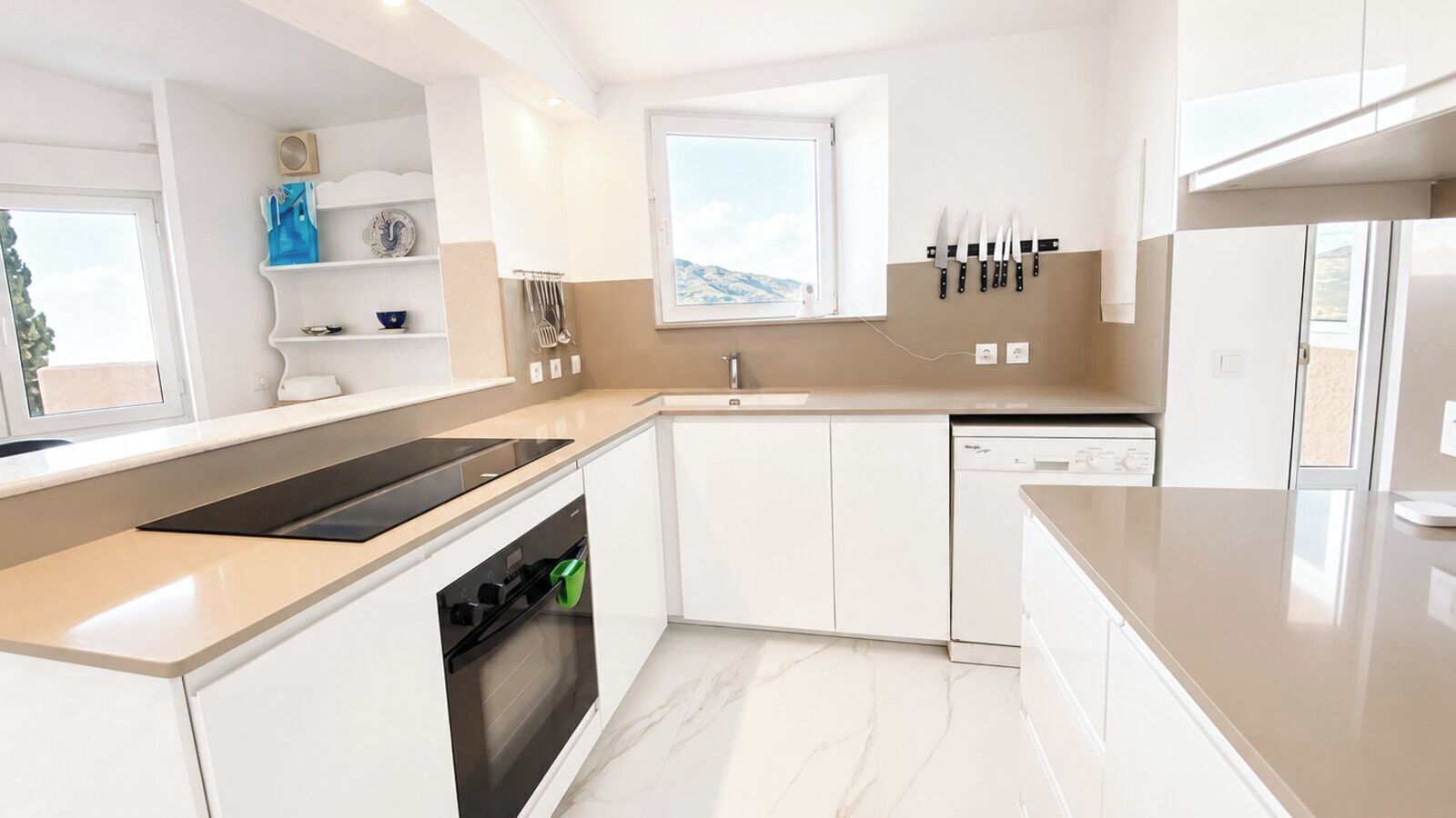 Una cocina moderna con armarios blancos, horno integrado, lavavajillas y ventana con vistas a las montañas.