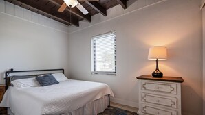 Een slaapkamer met een bed, nachtkastje, kapstok, raam met jaloezieën en een plafondventilator.