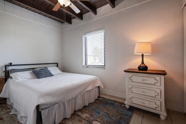 A bedroom with a bed, a nightstand, a dresser, a window with blinds, and a ceiling fan.