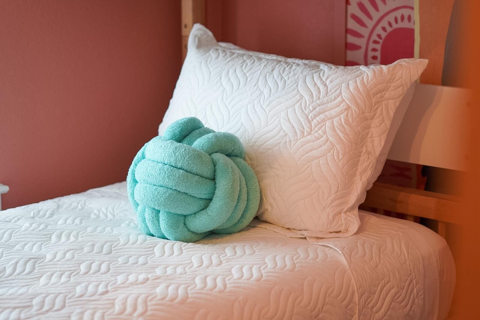 A bed with a white quilted headboard and a single teal-colored, braided pillow.