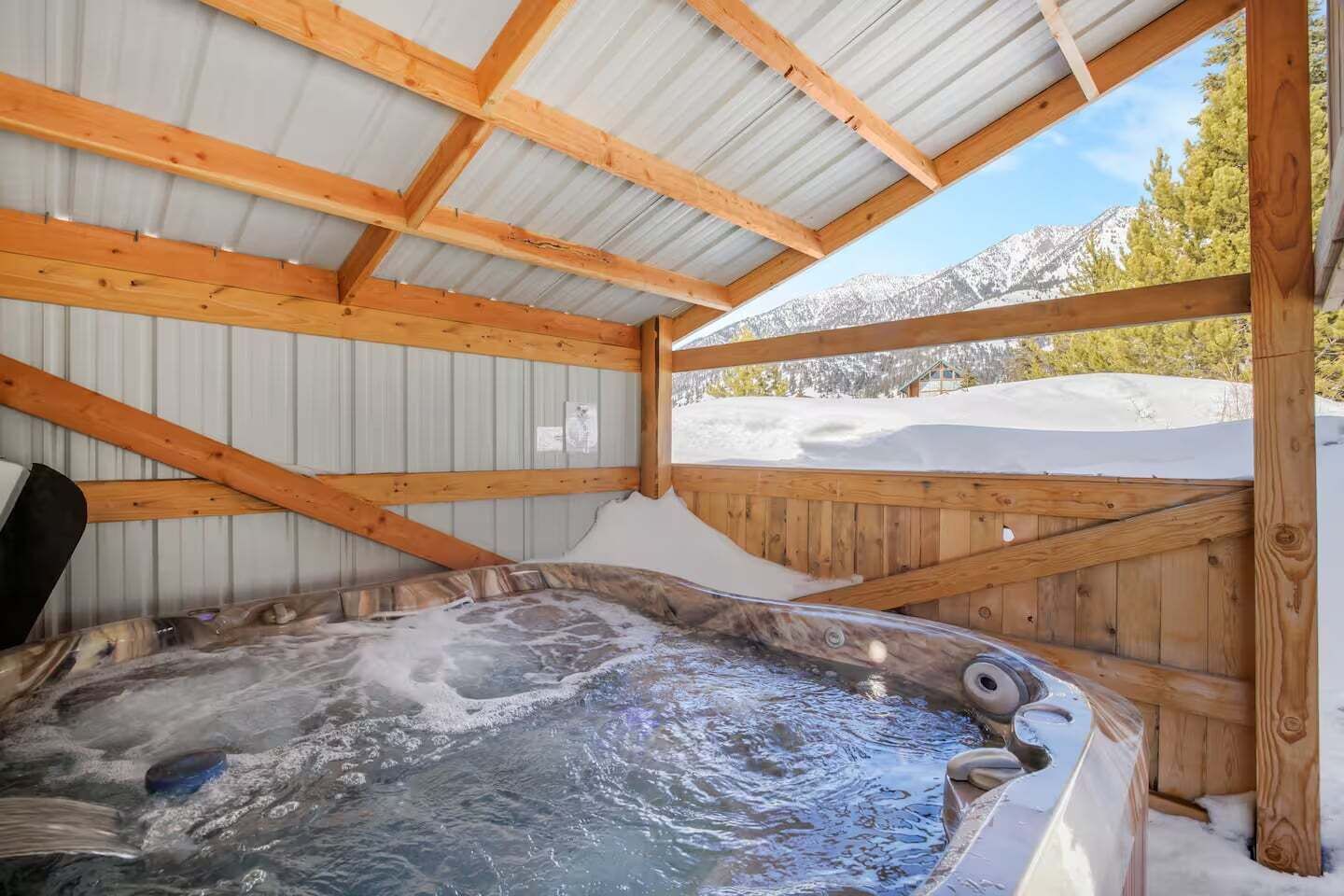A hot tub with a wooden deck and metal roof, surrounded by snow-covered mountains.