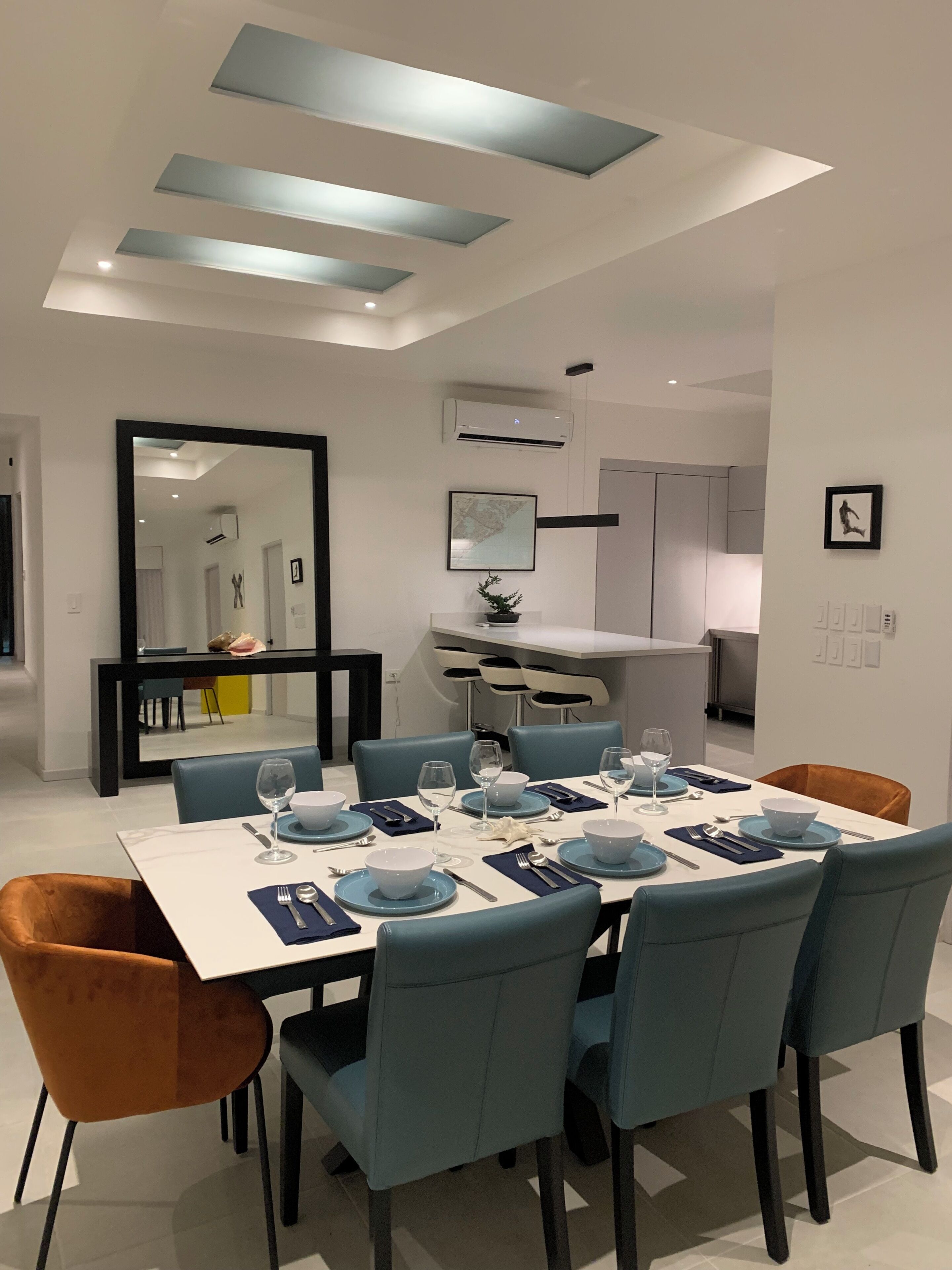 A modern dining area with a white table set for six, blue chairs, and a black-framed mirror on the wall.