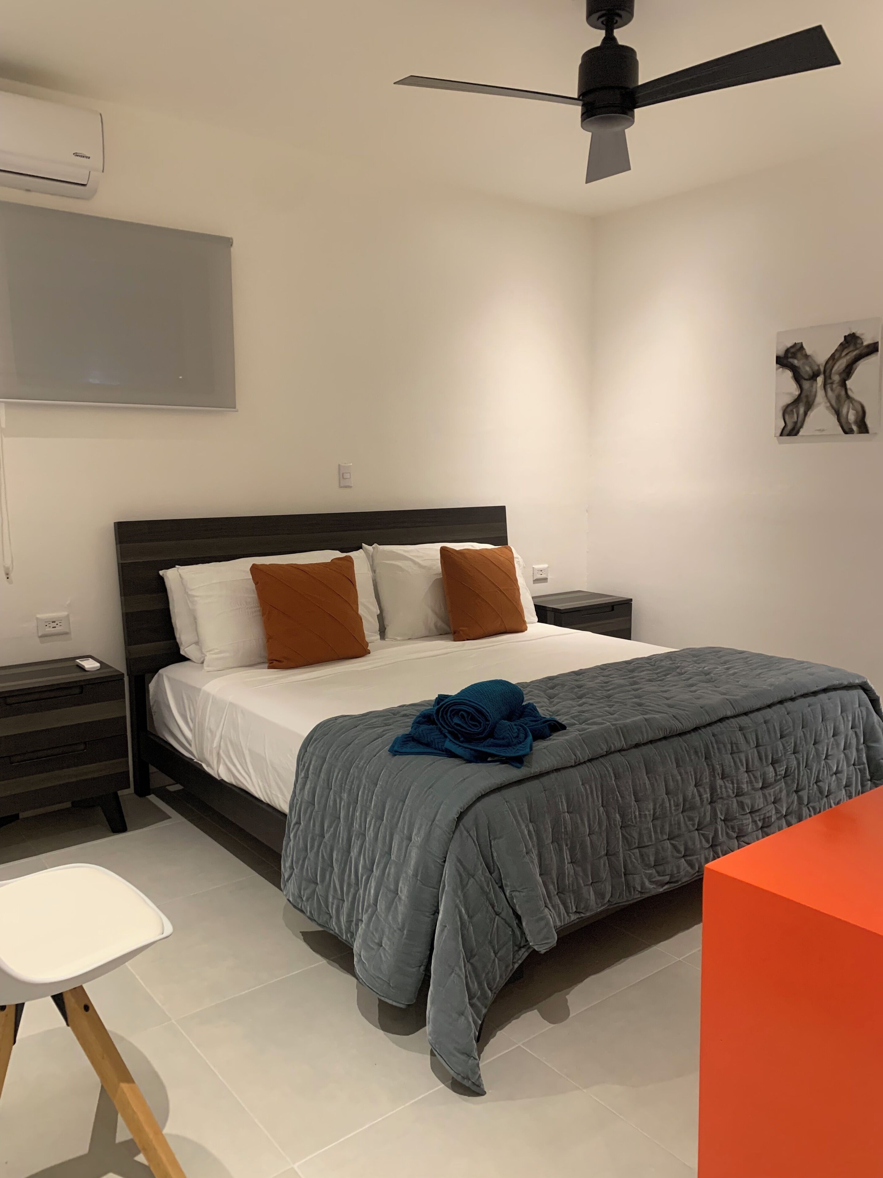 A bedroom with a bed, a nightstand, a chair, a ceiling fan, and a painting on the wall.