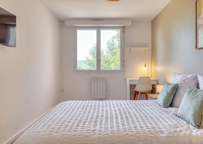 Ein Schlafzimmer mit einem Bett, einem Schreibtisch und einem Stuhl. Ein Fenster bietet Blick auf Bäume.