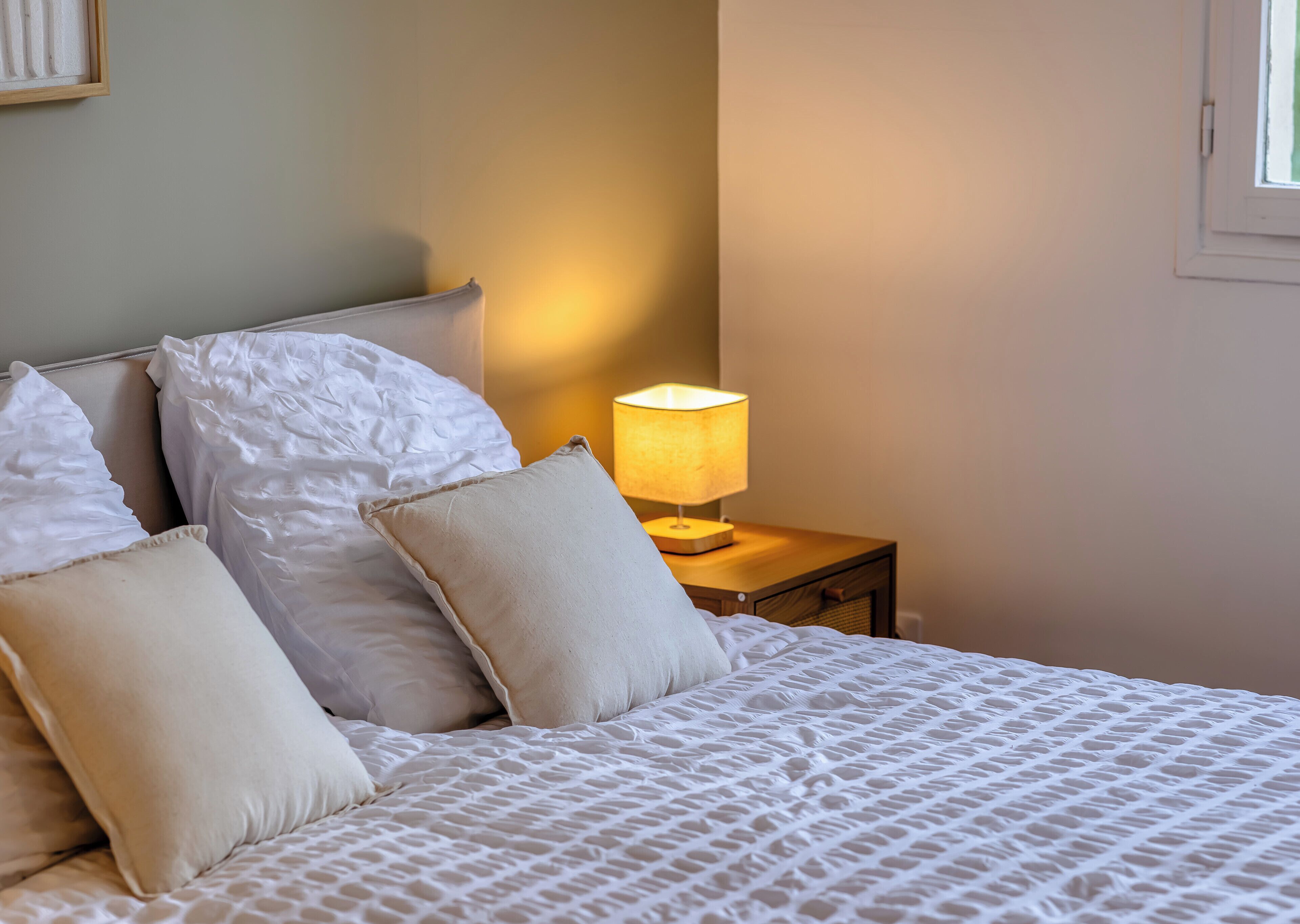 A neatly made bed with white linens and pillows, a bedside lamp, and a nightstand.