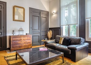 A living room with a leather sofa, a wooden dresser, a glass coffee table, and a large window providing a view of trees. - Valeria - Souplex élégant avec parking proche gare Rouen (Rouen)