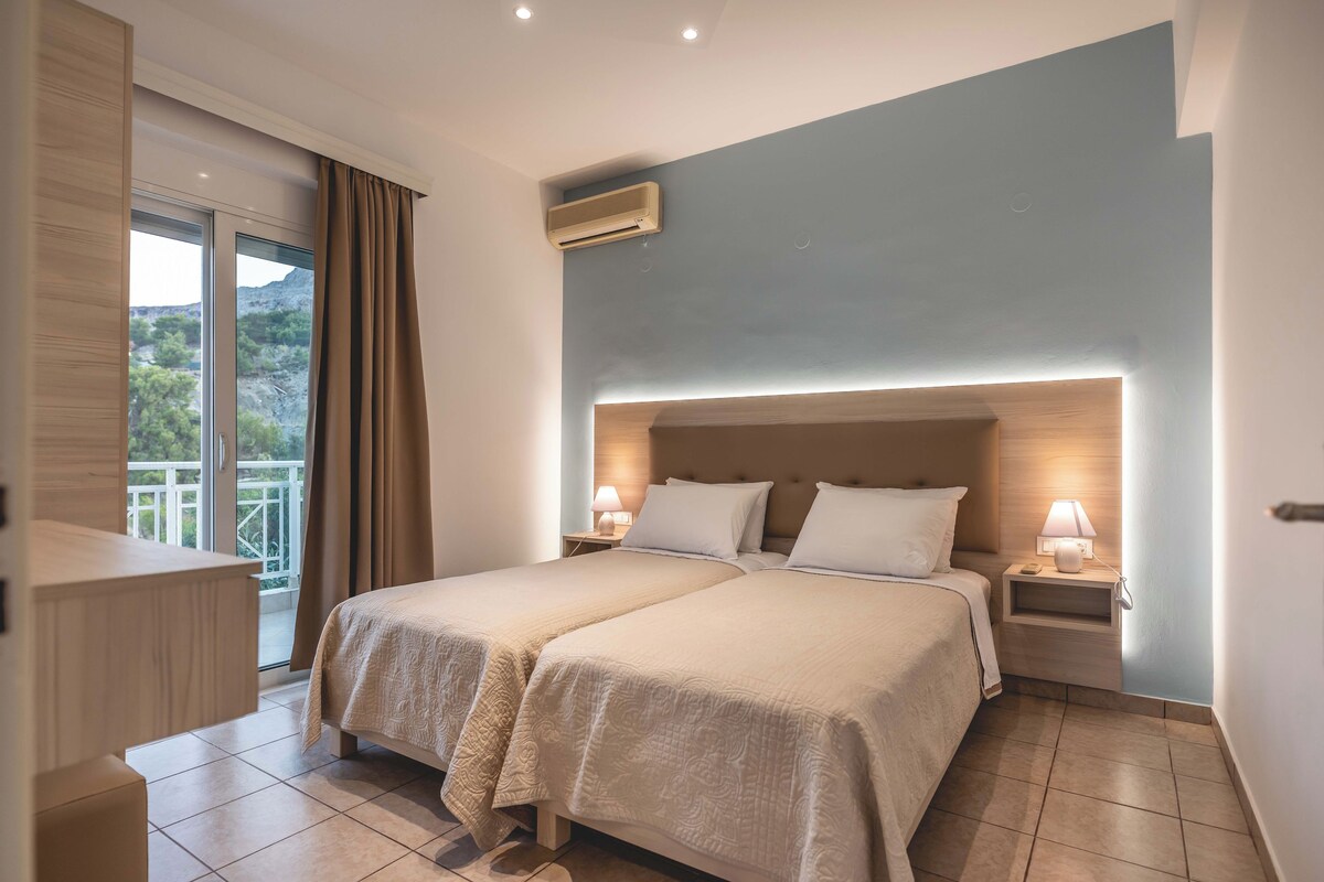 Una camera d'albergo con un letto grande, due comodini, un tavolino e una finestra con tende, oltre a un condizionatore d'aria.