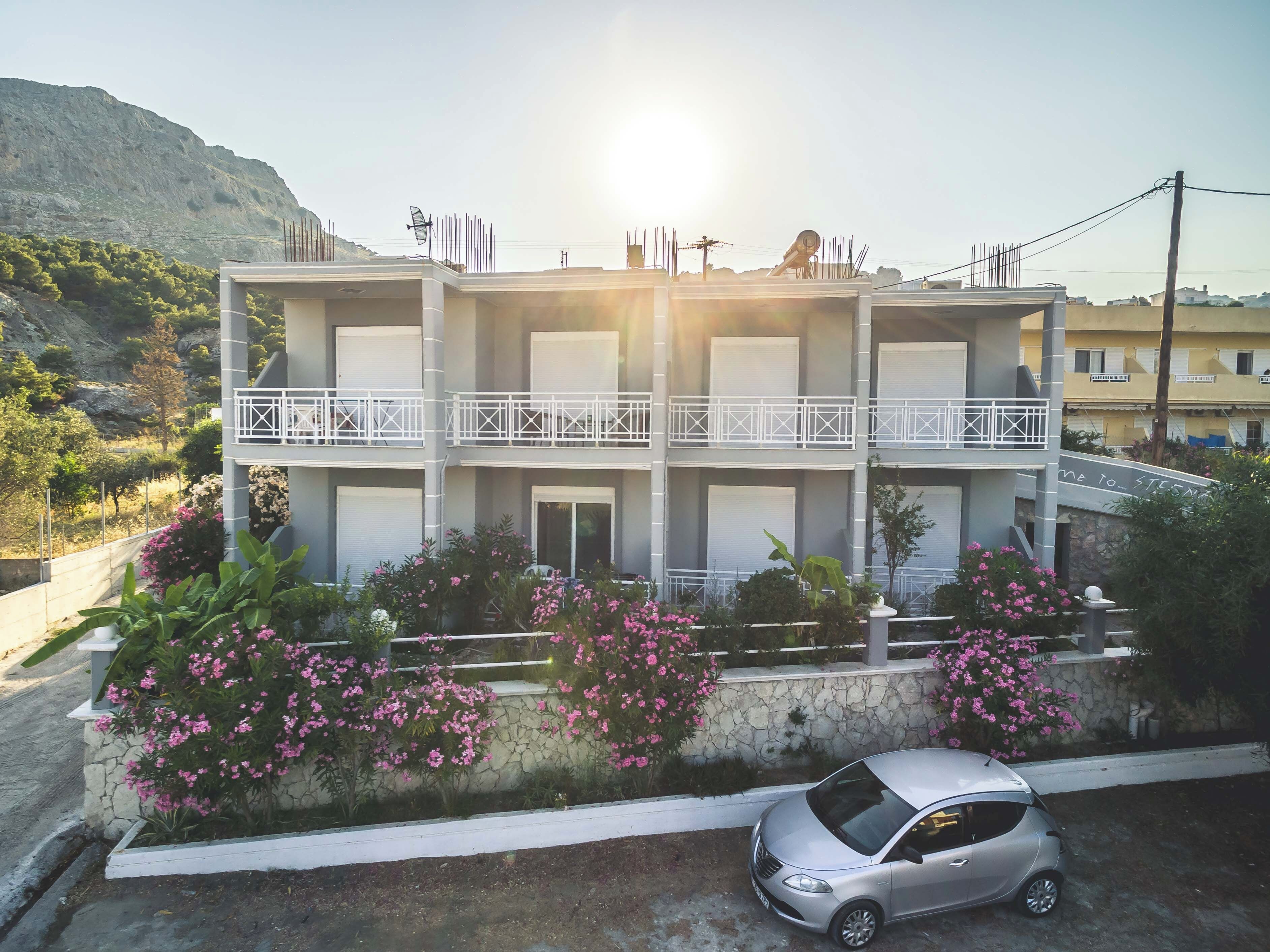 Un immeuble à plusieurs étages doté de balcons, une voiture garée devant, et un paysage montagneux en arrière-plan.