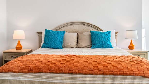 En snyggt bäddad säng med ett orangefärgat överkast, två blå kuddar och en beige sänggavel. Det står två sängbord bredvid sängen, med lampor och en liten låda i varje.