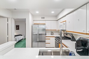 A compact kitchen with white cabinets, a stainless steel refrigerator, a microwave, and a stove. - Skinny Dippin 2 Bedroom Townhouse (Palm Springs)