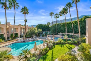 A pool area with lounge chairs and umbrellas, surrounded by palm trees and greenery, with a mountain backdrop. - Skinny Dippin 2 Bedroom Townhouse (Palm Springs)