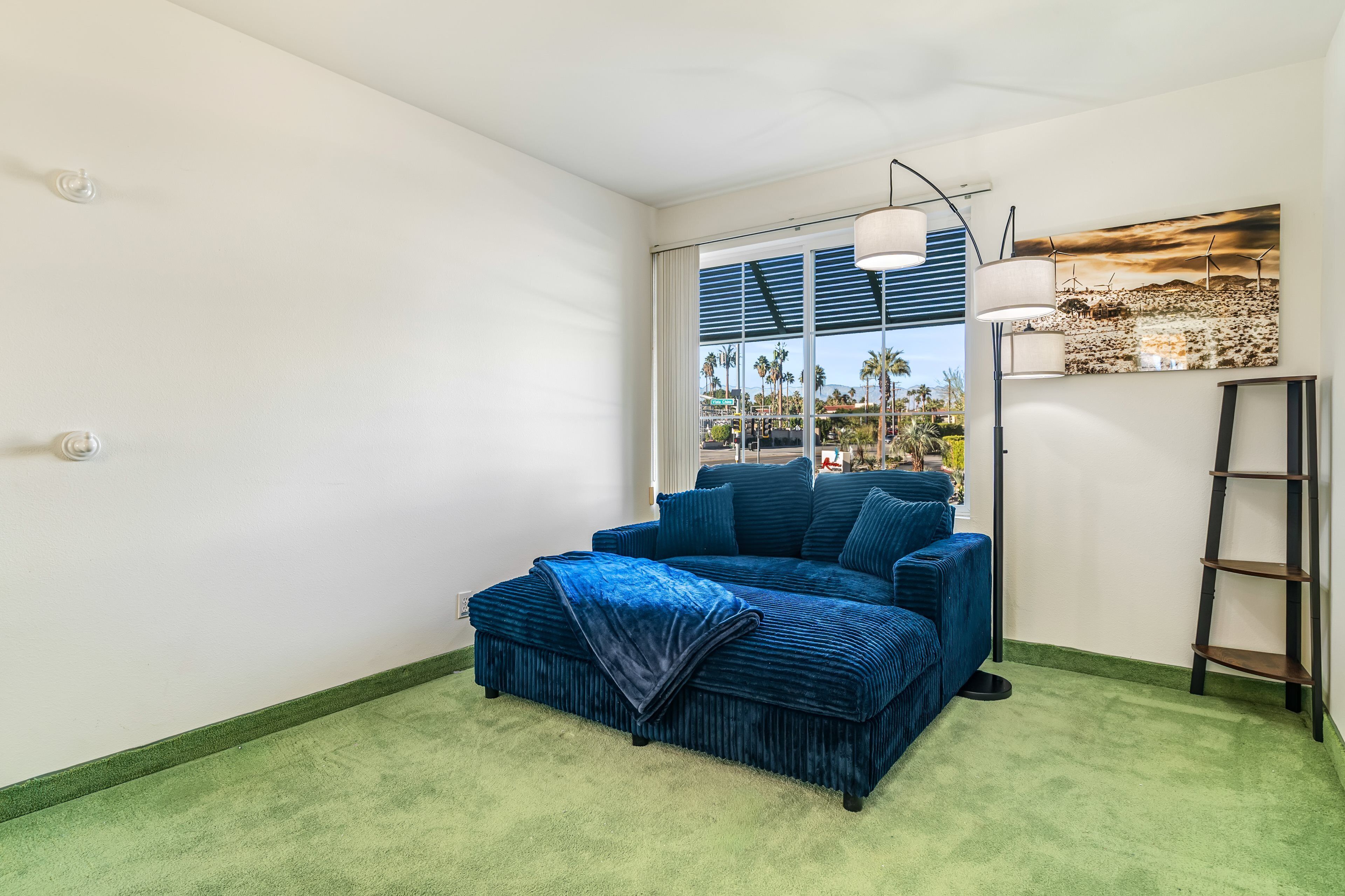 A modern living room with a blue sofa, a green carpet, a large window with blinds, a floor lamp, and a painting on the wall.