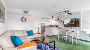 Ett modernt vardagsrum med en beige soffa, blå och orange kuddar, ett soffbord med blommor, en matplats med ett vitt bord och stolar samt ett kök i bakgrunden.
