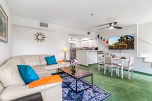 A modern living room with a beige sofa, blue and orange cushions, a coffee table with flowers, a dining area with a white table and chairs, and a kitchen in the background. - Skinny Dippin 2 Bedroom Townhouse (Palm Springs)