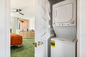 Ein Waschraum mit einer Frontlader-Waschmaschine und einem Trockner, einem Handtuchhalter und einer Tür zu einem Schlafzimmer mit Bett und Kommode.