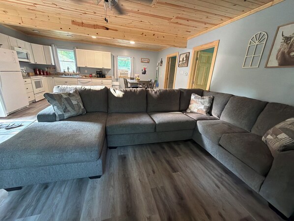 A modern living room with a sectional sofa, wooden flooring, and a kitchen in the background.