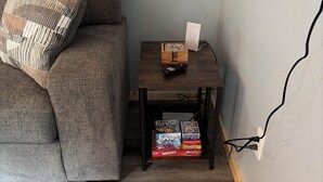 Una habitación con un sofá gris, una pequeña mesita de madera con un control remoto y una caja de juegos, y suelo de madera.