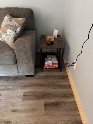 A room with a grey sofa, a small wooden side table with a remote control and a box of games, and a wooden floor.